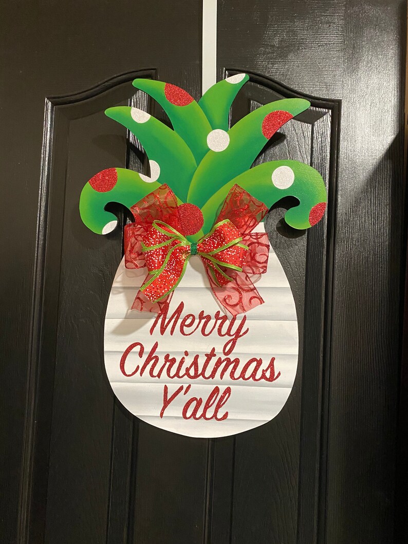 K&ouml;nnte beinhalten: Ein wei&szlig;er, ananasf&ouml;rmiger T&uuml;rkranz mit dem Text "Merry Christmas Y'all" in rotem Glitzer. Die Ananas hat gr&uuml;ne Bl&auml;tter mit roten und wei&szlig;en Punkten und eine rote Schleife mit gr&uuml;nem Besatz.