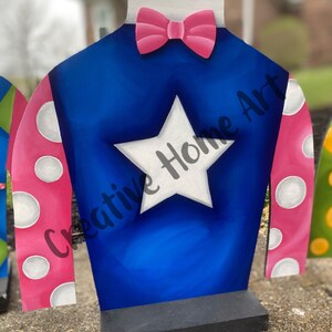 Puede incluir: Una pieza decorativa de madera con forma de casaca de jockey. La casaca es azul con una estrella blanca, mangas rosas con lunares blancos y una pajarita rosa. El texto "Creative Home Art" es visible en la casaca.