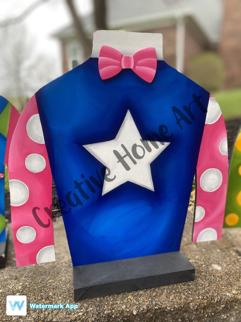 Puede incluir: Una pieza decorativa de madera con forma de casaca de jockey. La casaca es azul con una estrella blanca, mangas rosas con lunares blancos y una pajarita rosa. El texto "Creative Home Art" es visible en la casaca.