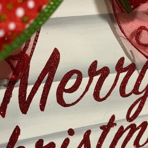 K&ouml;nnte beinhalten: Ein wei&szlig;es Holzschild mit den Worten "Merry Christmas" in rotem Glitzer.