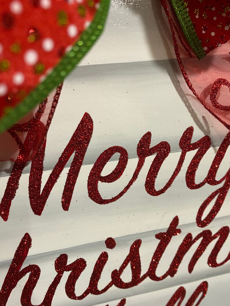 K&ouml;nnte beinhalten: Ein wei&szlig;es Holzschild mit den Worten "Merry Christmas" in rotem Glitzer.