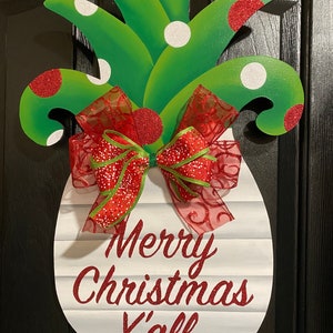 K&ouml;nnte beinhalten: Ein wei&szlig;er, ananasf&ouml;rmiger T&uuml;rkranz mit einer roten und gr&uuml;nen Schleife. Die Ananas hat gr&uuml;ne Bl&auml;tter mit wei&szlig;en und roten Punkten. Der Kranz tr&auml;gt den Text "Merry Christmas Y'all" in rotem Glitzer.
