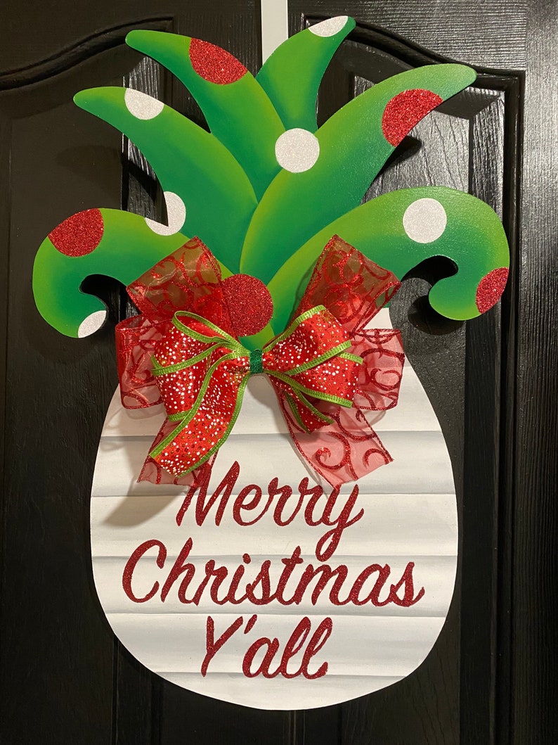 K&ouml;nnte beinhalten: Ein wei&szlig;er, ananasf&ouml;rmiger T&uuml;rkranz mit einer roten und gr&uuml;nen Schleife. Die Ananas hat gr&uuml;ne Bl&auml;tter mit wei&szlig;en und roten Punkten. Der Kranz tr&auml;gt den Text "Merry Christmas Y'all" in rotem Glitzer.