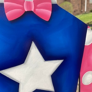 Puede incluir: Un recorte de madera de tem&aacute;tica patri&oacute;tica con una estrella blanca sobre un fondo azul, una pajarita rosa y un cuello blanco. El dise&ntilde;o incluye una secci&oacute;n de lunares rosas y blancos. La obra de arte est&aacute; pintada con un acabado brillante.