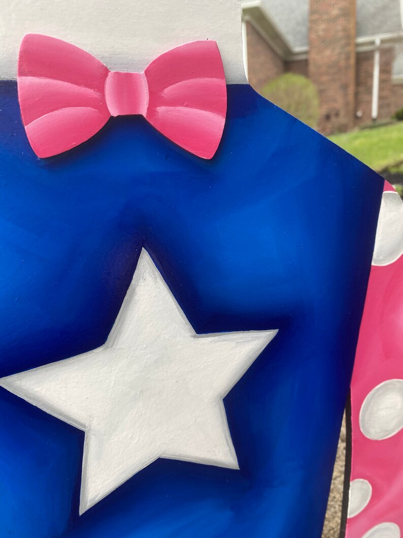 Puede incluir: Un recorte de madera de tem&aacute;tica patri&oacute;tica con una estrella blanca sobre un fondo azul, una pajarita rosa y un cuello blanco. El dise&ntilde;o incluye una secci&oacute;n de lunares rosas y blancos. La obra de arte est&aacute; pintada con un acabado brillante.