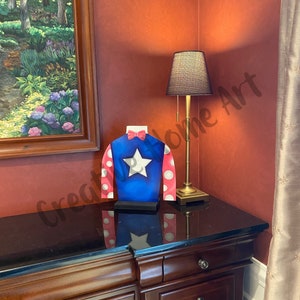 Puede incluir: Una pieza decorativa azul, roja y blanca con un dise&ntilde;o de estrella. La pieza se exhibe sobre una superficie de madera oscura, junto a una l&aacute;mpara dorada con una pantalla negra. Una pintura de un paisaje est&aacute; en el fondo.
