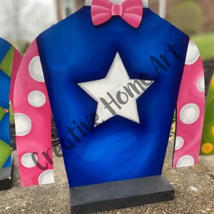Puede incluir: Recorte de madera de la casaca de un jockey. La casaca es azul con una estrella blanca, mangas rosas con lunares blancos y una pajarita rosa. El texto "Creative Home Art" es visible.