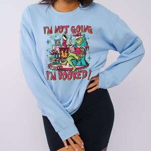 Peut inclure: Sweat-shirt bleu clair avec le texte "I'M NOT GOING I'M BOOKED !" et un dessin d'un personnage vert avec un chapeau de Père Noël lisant un livre. Le dessin comprend une cheminée, des cadeaux et une chaussette de Noël.