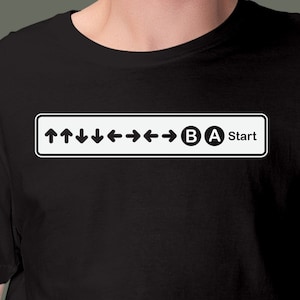 May include: Black t-shirt with a white rectangular graphic featuring directional arrows, the letters "B" and "A", and the word "Start". The design is a reference to a classic video game controller input sequence.