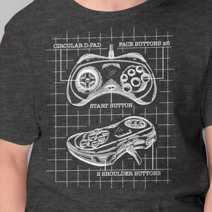 May include: A dark heather grey t-shirt with a white graphic of a video game controller. The graphic shows the controller's layout with labels for the circular d-pad, face buttons, start button, and shoulder buttons.
