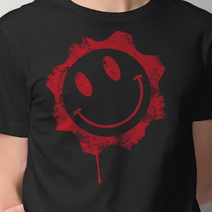 Puede incluir: Camiseta negra con un gráfico de cara sonriente roja y desgastada dentro de una forma de engranaje. El gráfico tiene un efecto de goteo.
