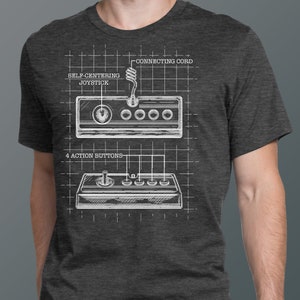 May include: A gray t-shirt with a white graphic of a vintage video game controller. The graphic includes the text "Self-Centering Joystick", "Connecting Cord", and "4 Action Buttons".