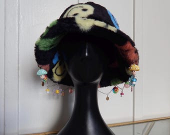 Könnte beinhalten: Ein schwarzer, flauschiger Bucket-Hut mit einem bunten psychedelischen Muster aus Smiley-Gesichtern, Blumen und Pilzen. Der Hut hat baumelnde Charms mit Pilzen, Blumen und Perlen.