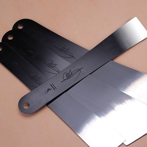 May include: Four black metal leather working tools with a polished metal edge. The tools have a hole at the top and a signature on the side.