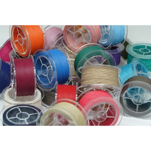 Puede incluir: Una colección de carretes de cordón encerado de colores, cada uno enrollado alrededor de un carrete de plástico transparente. Los cordones son de varios colores, incluidos rojo, azul, verde, marrón y beige.