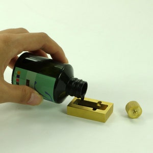 Puede incluir: Una mano vierte tinta negra de una botella en un tintero de lat&oacute;n con un peque&ntilde;o pomo de lat&oacute;n en el lateral.