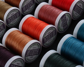 Op de afbeelding: Close-up van spoelen met gekleurd polyester garen, elk met het label "Ten Colors of Light" en "0,4 mm". De spoelen zijn in een halve cirkel op een zwart oppervlak gerangschikt.
