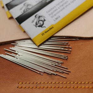May include: A close-up of a pile of 25 silver needles on a brown leather surface. The needles are arranged in a fan shape. The needles are for leather stitching. A yellow and white package with the text "Saddlers' Needles Aiguilles à  cuir Straminadele  cuir L1912 25 Needles" is visible in the background.