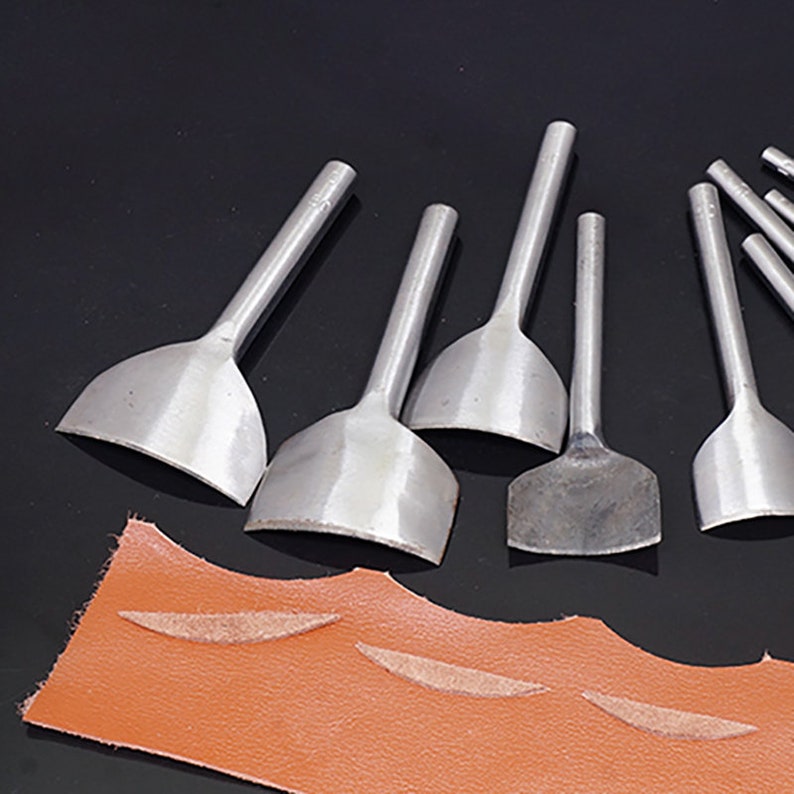 Puede incluir: Un conjunto de herramientas de trabajo de cuero de metal plateado con cuchillas curvas y mangos largos. Las herramientas est&aacute;n dispuestas sobre una superficie oscura cerca de un trozo de cuero marr&oacute;n con marcas de corte.