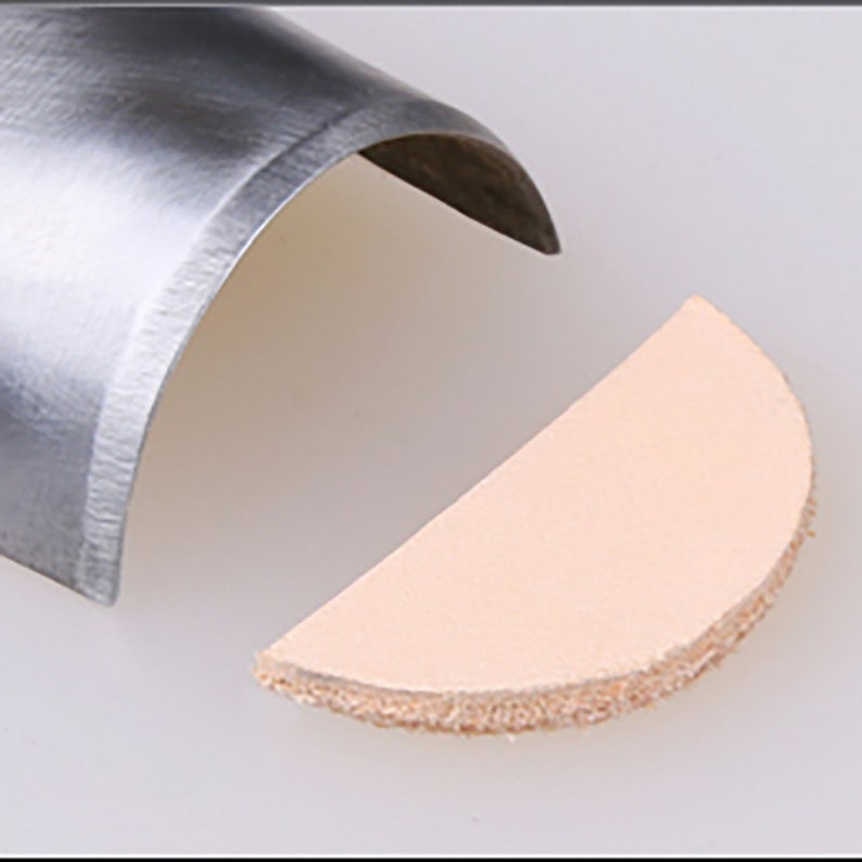 May include: A close-up of a metal leather punch tool with a half-circle cut out of a piece of light brown leather.