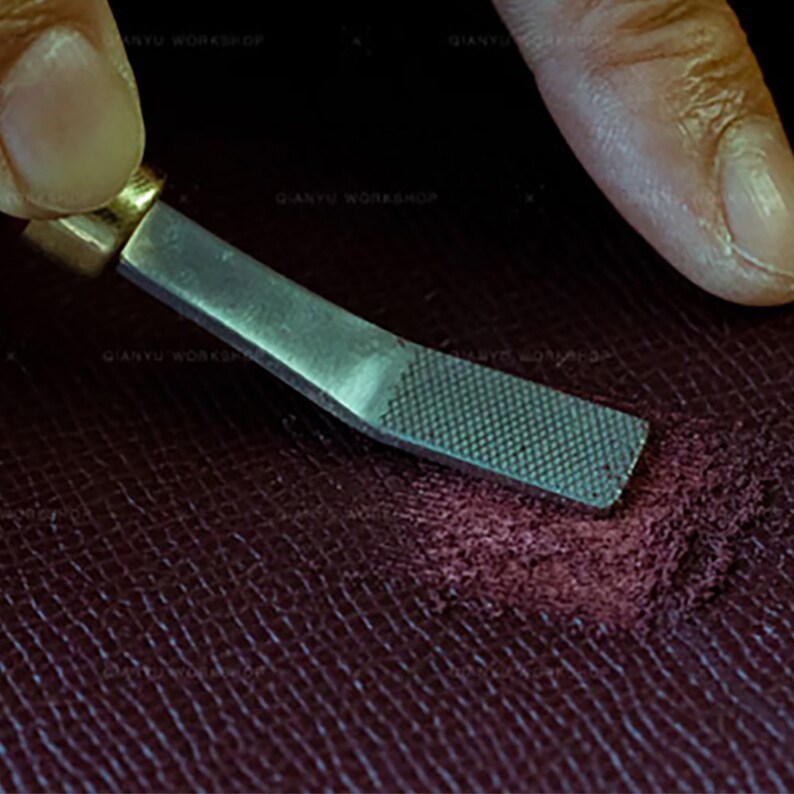 Peut inclure: Gros plan d'une lime m&eacute;tallique utilis&eacute;e pour lisser un morceau de cuir bordeaux. La lime a une surface textur&eacute;e et un manche en laiton.