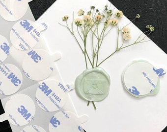 May include: A white card with a green wax seal and dried white flowers. The wax seal has a small envelope design. There are several 3M double-coated tissue tape circles around the card.