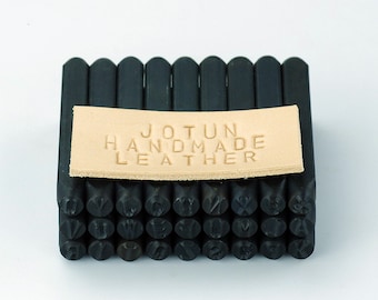 Könnte beinhalten: Ein Satz schwarzer Metallbuchstaben-Stempel, die in einem Stapel angeordnet sind. Die Stempel werden verwendet, um individuelle Beschriftungen auf Leder zu erstellen. Ein kleines braunes Leder-Etikett mit dem Text "JOTUN HANDMADE LEATHER" ist oben auf den Stempeln platziert.