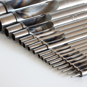 May include: A collection of metal hole punch tools, each with a different diameter, arranged on a white surface. The tools are cylindrical with a hollow centre and a sharpened edge. The tools are made of a shiny, silver-coloured metal.