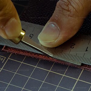 Peut inclure: Une personne utilise un petit outil en m&eacute;tal pour couper un morceau de cuir brun. L'outil a une poign&eacute;e en laiton et une lame dentel&eacute;e. Le cuir est coup&eacute; sur un tapis de coupe noir avec un motif quadrill&eacute;.