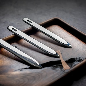May include: Three silver metal tools with pointed ends and a small loop at the top, arranged on a wooden tray.
