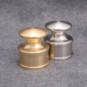 May include: Two metal bottle stoppers. One is gold-toned, the other is silver-toned. Each stopper has a cylindrical base and a rounded top. The stoppers are displayed on a grey surface.