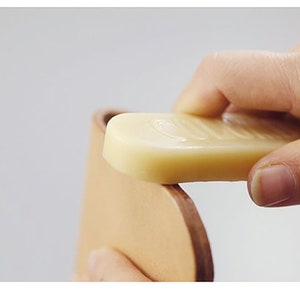 May include: A hand rubs a bar of light beige leather conditioner onto a piece of light brown leather.