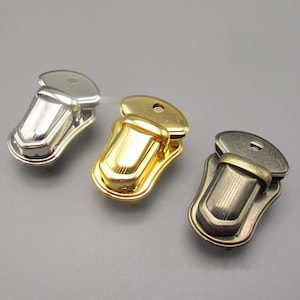 Pode incluir: Três fechos de metal em acabamentos prata, ouro e bronze antigo. Cada fecho tem uma forma retangular decorativa ligeiramente curva com um pequeno furo na parte superior.