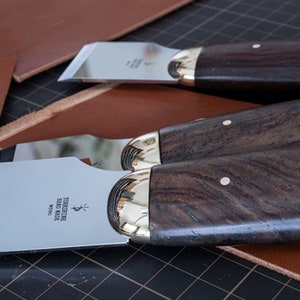 Op de afbeelding: Drie gereedschappen voor het bewerken van leer met houten handvatten en metalen bladen. De handvatten zijn van donkerbruin hout met gouden accenten. De bladen zijn zilverkleurig en hebben een lichte kromming. De gereedschappen zijn gemarkeerd met "Yorkshire Hand Made M300".