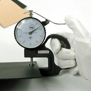May include: A black leather thickness gauge with a dial reading 0-10mm and 0.01mm. The gauge has a black handle and a silver metal probe. The gauge is being used to measure the thickness of a piece of black leather.