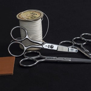 May include: A selection of silver sewing scissors, a spool of white thread, and a small piece of brown leather on a black background. The scissors have curved handles and sharp blades, suitable for crafting and sewing.