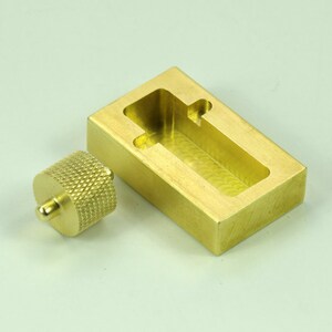 Puede incluir: Un molde de lat&oacute;n con forma rectangular y una peque&ntilde;a perilla redonda. El molde tiene una abertura rectangular con un borde redondeado.