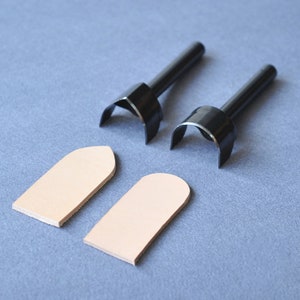 May include: Two black leathercraft tools with curved cutting edges and long handles, and two pieces of beige leather. The tools are for shaping leather, and the leather pieces are rectangular with rounded tops.
