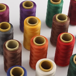 Puede incluir: Surtido de carretes de hilo de colores, incluyendo morado, verde, marrón, amarillo, naranja, rojo, gris, blanco y marrón. Los carretes están dispuestos en un patrón circular sobre una superficie blanca.