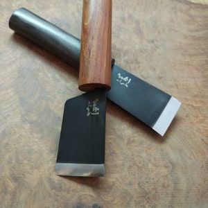 Puede incluir: Dos cuchillos de cocina japoneses con mangos de madera. Los cuchillos tienen hojas negras con detalles plateados y caracteres japoneses en las hojas.