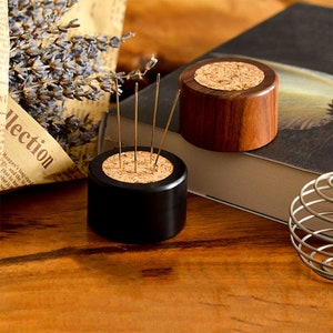 May include: Two wooden pincushions with cork tops. One is black and the other is brown. Both have needles sticking into the cork.