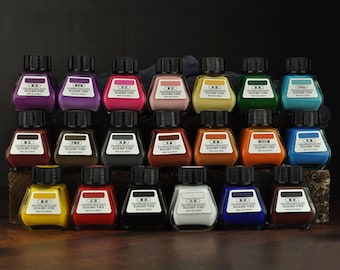 Puede incluir: Un conjunto de 24 pequeños frascos de vidrio con tapas negras, cada uno contiene una pasta de tinte para cuero de un color diferente. Los frascos están dispuestos en tres filas de ocho, con colores que van de claro a oscuro.
