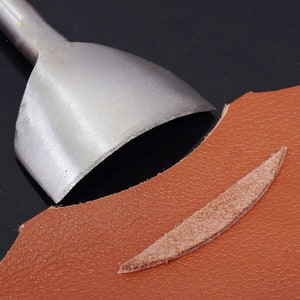 Puede incluir: Una herramienta de marroquiner&iacute;a met&aacute;lica con una cuchilla curva se coloca sobre un trozo de cuero marr&oacute;n. Se ve un trozo de cuero recortado, con forma de &oacute;valo largo y fino. La herramienta est&aacute; dise&ntilde;ada para cortar y dar forma al cuero.