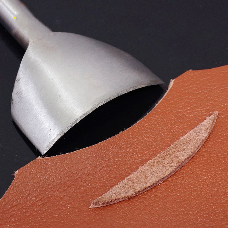 Puede incluir: Una herramienta de marroquiner&iacute;a met&aacute;lica con una cuchilla curva se coloca sobre un trozo de cuero marr&oacute;n. Se ve un trozo de cuero recortado, con forma de &oacute;valo largo y fino. La herramienta est&aacute; dise&ntilde;ada para cortar y dar forma al cuero.