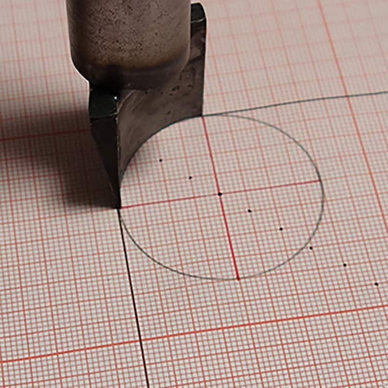 May include: A close-up of a metal tool used to cut a circle into a piece of paper. The tool is positioned over a circle drawn on a gridded sheet of paper.