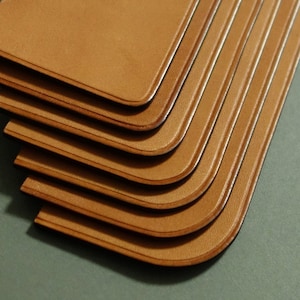 May include: A stack of six brown leather bookmarks with rounded edges. The bookmarks are stacked on top of each other, with the top bookmark being the most visible.