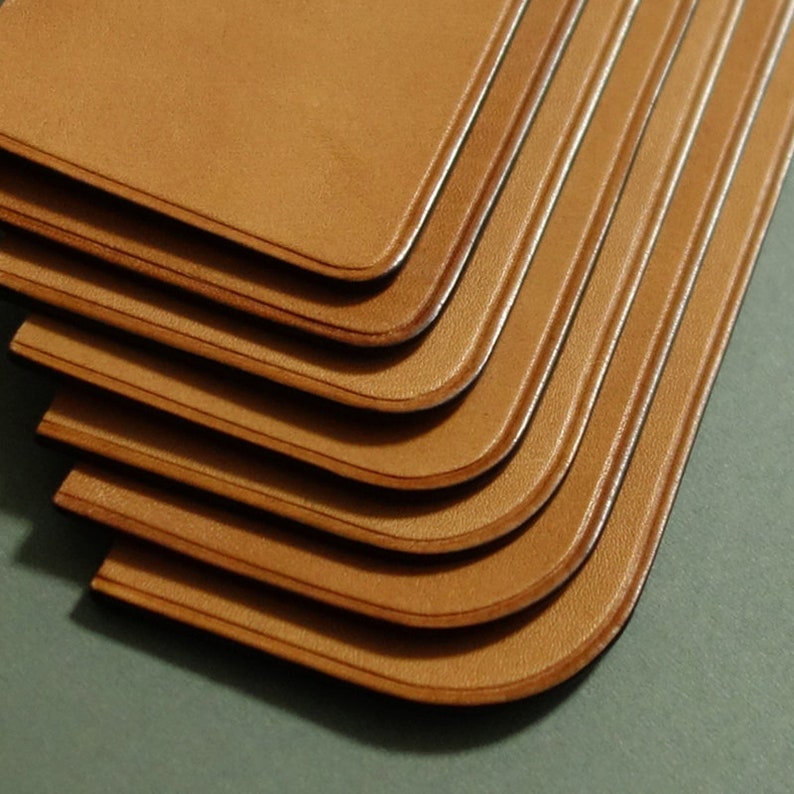 May include: A stack of six brown leather bookmarks with rounded edges. The bookmarks are stacked on top of each other, with the top bookmark being the most visible.