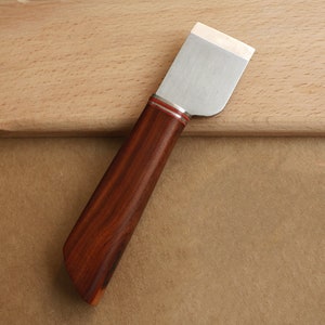 Puede incluir: Un pequeño cuchillo con mango de madera marrón y hoja de plata. La hoja está ligeramente curvada y tiene una punta redondeada.