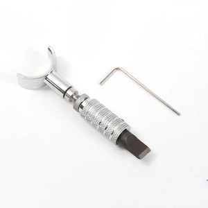 May include: A silver-coloured swivel knife for leather crafting featuring a U-shaped yoke, a textured cylindrical handle for grip, and a flat blade tip. An L-shaped metal hex key is included alongside the tool for adjustment and maintenance.