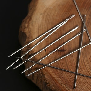 May include: A set of six silver sewing needles on a wooden surface.